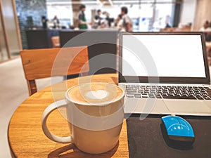 A white cup hot capuccino with white heart mug on top, a white screen computer labtop and blue mouse on brown wooden table