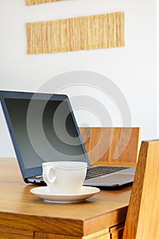 White cup and laptop on natural wood dining table. Remote work c