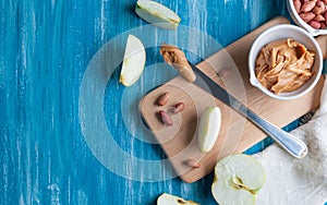 White cup with peanut butter. On a wooden board. Nearby sliced â€‹â€‹apple, peanuts and a table knife with pasta..Flat lay