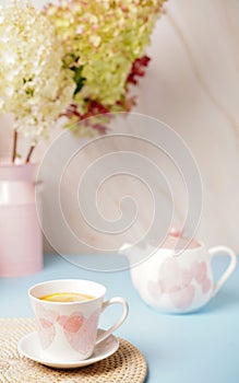 white cup with pink hearts a teapot and a bouquet of flowers on beige-blue background. Breakfast, morning tea