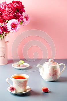 white cup with pink hearts a teapot, sweets and a bouquet of flowers on a pink-gray background. Breakfast, morning tea