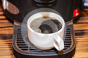 A white cup with prepared black coffee is in the coffee machine. Breakfasts and business meetings. Close-up