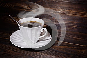 White cup and saucer with coffee and spoon on brown wooden surface, steam over the cup.