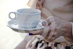 A white cup with a saucer in the graceful hand of a woman in beige clothes