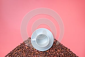 A white cup on a saucer stands on a hill of coffee beans on a pink background