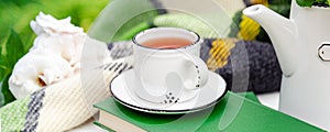 White cup of tea on books with green warm plaid, spring flowers on white table outdoor. Hot drink at breakfast in garden. Long web