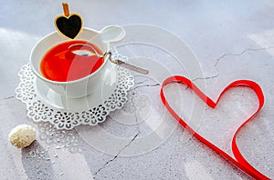A white cup of tea on a delicate white saucer with a small black heart on a cup and with the original curved spoon on