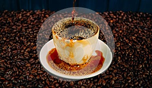 A white cup in which coffee is poured stands on a table covered with roasted coffee beans