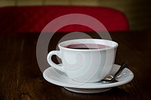 A white cup on a white saucer and spoon, with red tea, on a dark wooden table with red chair in a dark cafe