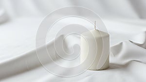A white cylindrical candle is placed on a soft white fabric surface