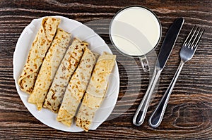 White dish with pancake rolls, cup of milk, knife and fork on table. Top view