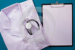 A white doctor`s uniform and a stethoscope in her pocket lie on the table, next to a notepad