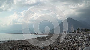 A white dog walking along the pebbly shore. Fishermen on the coast. Coast with mountains and cloudy sky in the background.