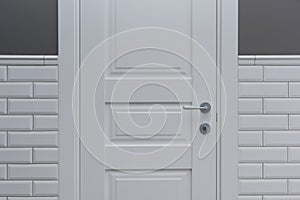 White door in the bathroom toilet room. Background gray painted wall covered with decorative ceramic tiles with white glossy brick