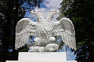 White doubleheaded metal eagle, looking in different directions, against the background of trees and blue sky with clouds