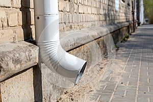 White downpipe on the wall of a brick building in dry, sunny weather. Tube, gutters. Drainpipe and asphalt sidewalk