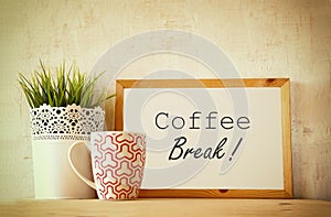 White drawing board with the phrase coffee break over wooden table with coffe cup and flower pot decoration . filtered image