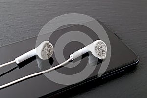 White earphones reflects in a black smartphone screen on a black stone surface. Modern personal device with touchscreen for music