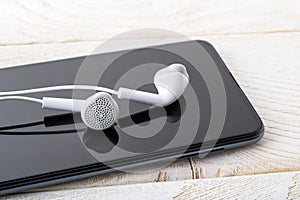 White earphones reflects in a black smartphone screen on a white wooden table. Listen to music. Modern personal device with