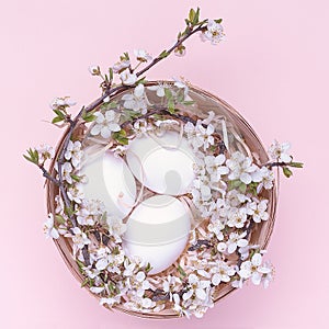 White easter eggs in a basket with flowers on a pink background. Square photograph. Top view