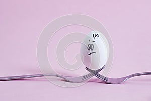 White egg with painted fase lies on crossed forks as an egg stand on a pink background