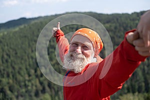 a white elderly man takes a selfie on the top of a mountain, against the background of the sky and the forest from a