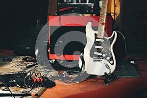 A white electric guitar and a red amplifier onstage with electrical audio cables and sound effects pedals