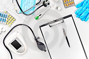 White electric tonometer with stethoscope, multicolored pills, mask and thermometer on white table. Top view