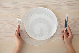 White empty plate and female hands holding silver fork and knife on wooden table background, top view