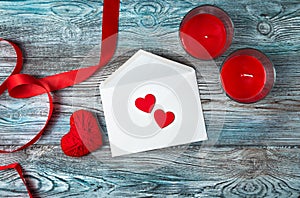 White envelope, red hearts, ribbon and candles on a wooden gray-blue background.