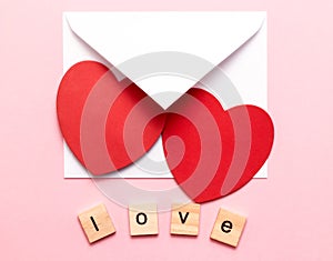 A white envelope with two red hearts and wooden cubes with the word Love on a pink background. A love letter