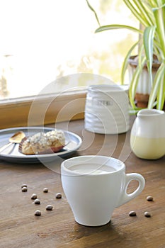 white espresso coffee cup, fresh baked eclair sweet dessert on plate kitchen table against window, utensils dishware