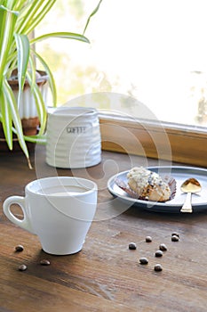 white espresso coffee cup, fresh baked eclair sweet dessert on plate kitchen table against window, utensils dishware