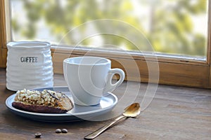 White espresso coffee cup, fresh baked eclair sweet dessert on plate kitchen table against window, utensils dishware, milk jug,