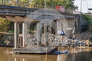 A white excavator works under a bridge over the Arno river to reinforce the embankment and structure, Calcinaia, Italy