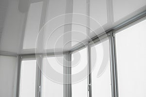 White fabric roller blinds on the plastic window on a balcony in the living room with a reflection in a stretch ceiling.