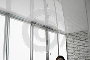 White fabric roller blinds on the plastic window on a balcony in the living room with a reflection in a stretch ceiling.