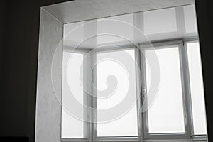 White fabric roller blinds on the plastic window on a balcony in the living room with a reflection in a stretch ceiling.