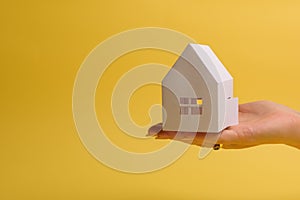 White family paper house in man hand on yellow background paper. Minimalistic style. Copy space. View from above. Horizontal