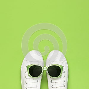 White female fashion sneakers and trendy sunglasses on green background. Flat lay top view copy space. Women`s shoes, accessories