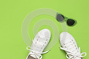 White female fashion sneakers and trendy sunglasses on green background. Flat lay top view copy space. Women`s shoes, accessories