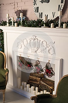 White fireplace with stucco patterns, candles, wach face, fir branches. Christmas decor. Close up. Selective focus