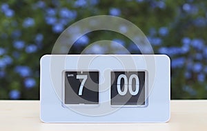 white flip clock showing 7.00 o clock on the table in the garden with purple flowers background