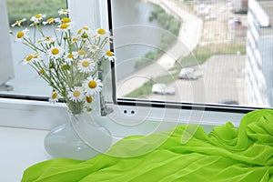 White flowers in a white vase on a white window-sill with bright green fabric and an open white window and street view with a rive