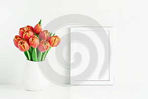 White frame mockup and red tulips in a white vase on a white table by the wall