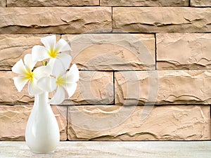 white frangipani or plumeria bouquet in pottery vase on shelf floor and beige sandstone brick wall with copy space