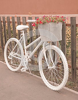 White ghost bike with a basket full of pink flowers parking next to a rustic wooden fence