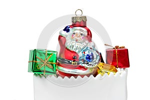 White gift bag filled with Santa Claus tree ornament and red, green and yellow foil wrapped presents.