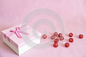 White gift box with pink lid and bow next to red metal balls on pink fabric background with copy space. Celebration concept