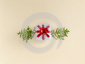 White gift box with red ribbon accompanied by arborvitae branches on beige background, horizontal.